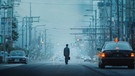Ein Mann mit einem kleinen Koffer läuft verloren und isoliert mitten auf der Fahrbahn einer Straße in einer japanischen Großstadt. | Bild: BR/Ossa Film/Abel van Erkel